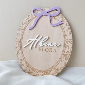 Pu&ograve; includere: Una targa ovale in legno con finitura a venature chiare e bordo floreale dorato. La targa mostra il nome Athena in lettere corsive bianche in rilievo sopra il nome ELORA in testo dorato, sormontata da un fiocco in acrilico color lavanda.