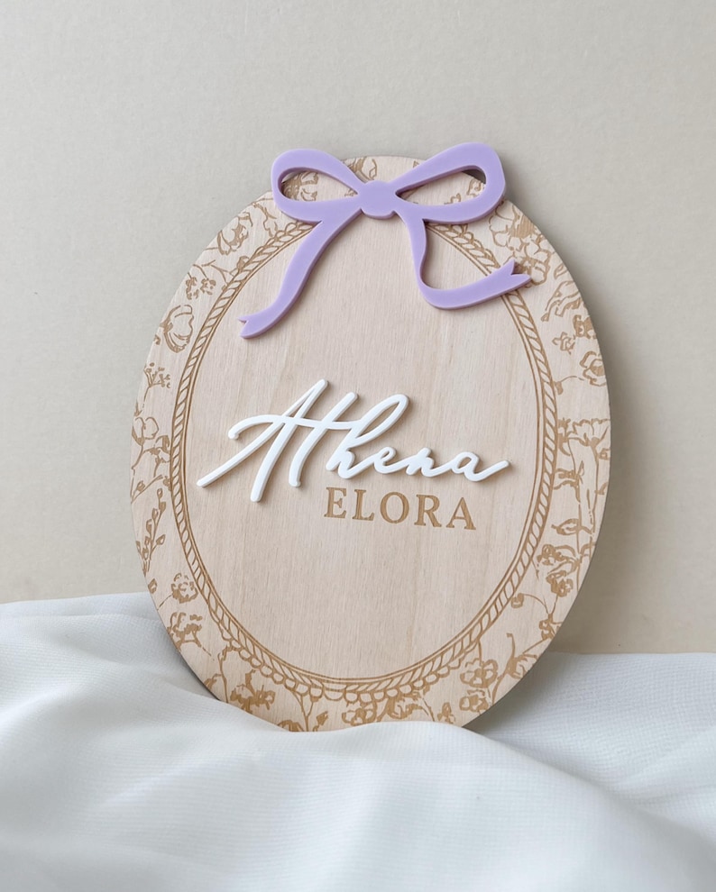Pu&ograve; includere: Una targa ovale in legno con finitura a venature chiare e bordo floreale dorato. La targa mostra il nome Athena in lettere corsive bianche in rilievo sopra il nome ELORA in testo dorato, sormontata da un fiocco in acrilico color lavanda.