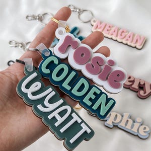 May include: A collection of layered acrylic keychains featuring custom names in various colors including white, dark green, teal, and dusty rose. Each keychain has a metal split ring attached with a clear plastic loop, displaying names like Wyatt, Colden, Rosie, and Meagan.