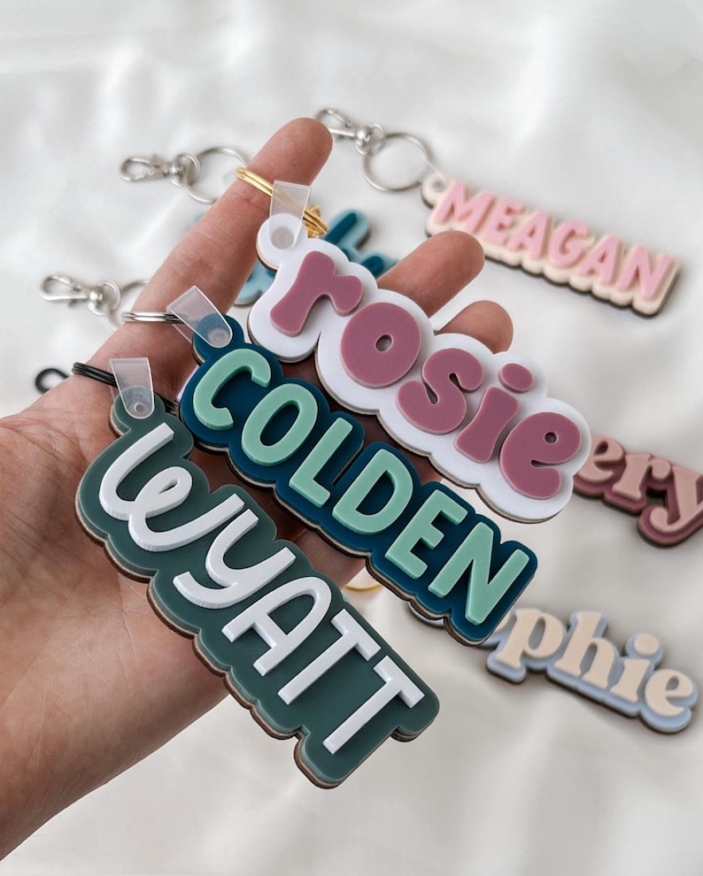 May include: A collection of layered acrylic keychains featuring custom names in various colors including white, dark green, teal, and dusty rose. Each keychain has a metal split ring attached with a clear plastic loop, displaying names like Wyatt, Colden, Rosie, and Meagan.