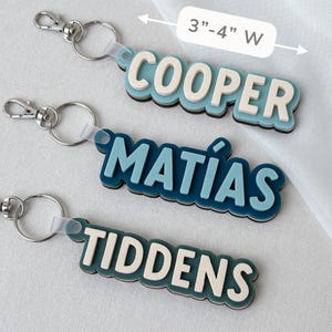 May include: Three custom keychains with metal lobster clasps and rings. Each features layered, raised lettering in blue and cream tones. The names COOPER, MAT&Iacute;AS, and TIDDENS are displayed. A label indicates a width of 3 inches to 4 inches.