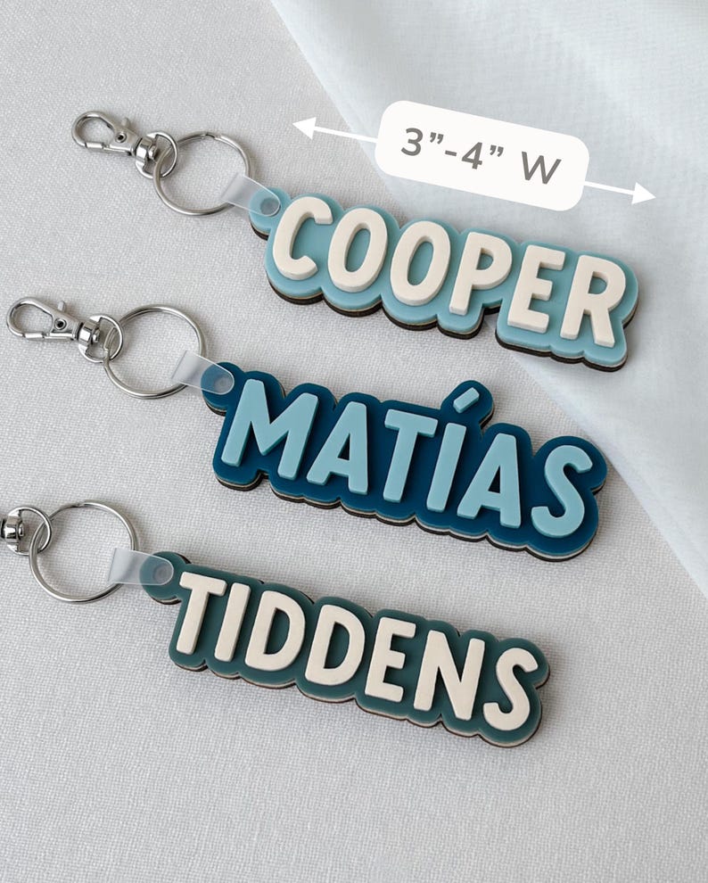 May include: Three custom keychains with metal lobster clasps and rings. Each features layered, raised lettering in blue and cream tones. The names COOPER, MAT&Iacute;AS, and TIDDENS are displayed. A label indicates a width of 3 inches to 4 inches.