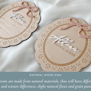 May include: Two oval wooden signs with scalloped edges and laser-cut floral patterns. Each sign features a raised dusty pink bow, white 3D script text reading "Allora", and the printed name "OLIVIA OCKER". One sign includes fields for date, weight, time, and length.