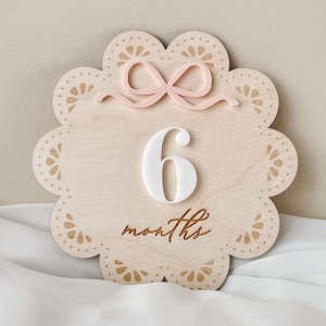 Crafted intricately from wood, the monthly milestone disc features a delicate bow and lace pattern, adding a charming touch to any baby girl's nursery.