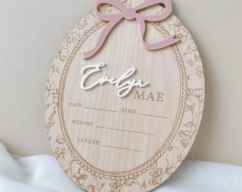 May include: An oval wooden birth announcement sign featuring a dusty pink acrylic bow at the top. The center displays the name Evelyn Mae in white and brown lettering, with engraved lines for date, time, weight, and length. A floral border surrounds the text.