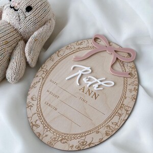 Pu&ograve; includere: Una targa nascita ovale in legno con un fiocco in acrilico rosa polvere, il nome Ruth Ann in corsivo bianco e campi incisi per data, peso, ora e lunghezza. Un coniglietto in maglia beige &egrave; posizionato accanto alla targa.