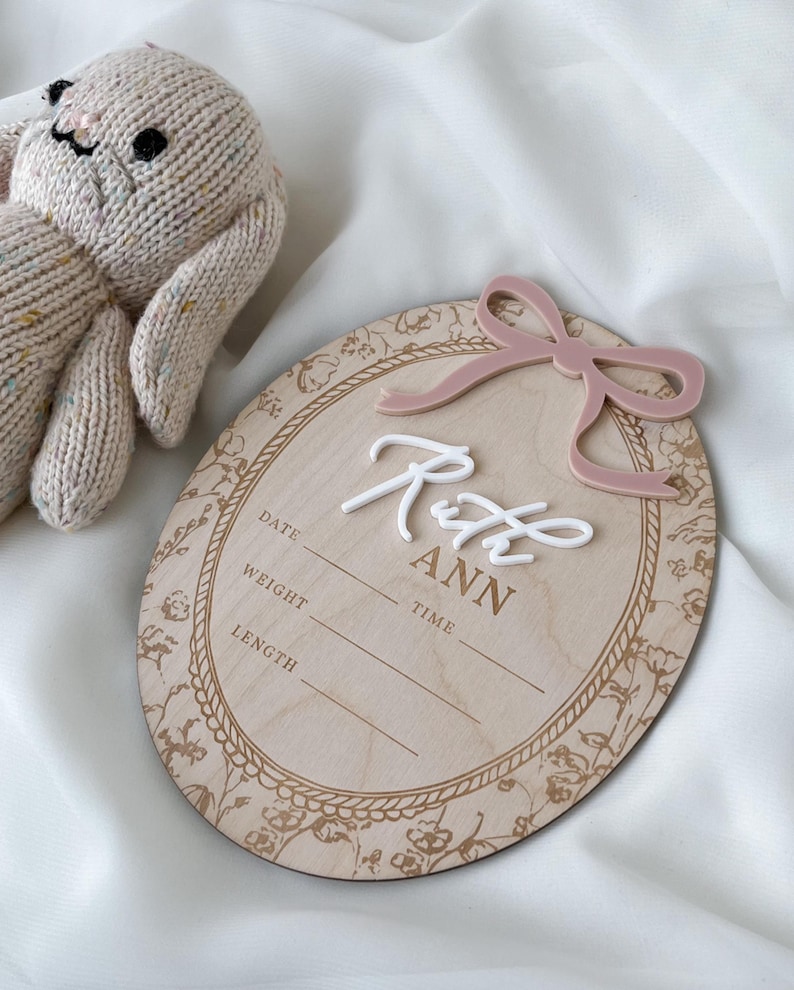Pu&ograve; includere: Una targa nascita ovale in legno con un fiocco in acrilico rosa polvere, il nome Ruth Ann in corsivo bianco e campi incisi per data, peso, ora e lunghezza. Un coniglietto in maglia beige &egrave; posizionato accanto alla targa.
