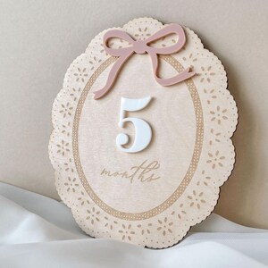 May include: An oval wooden milestone plaque featuring a laser-cut lace-style border, a dusty pink acrylic bow at the top, and a white acrylic number 5 centered above the engraved cursive text months, all set against a light wood grain background.