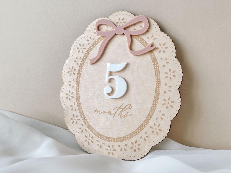 May include: An oval wooden milestone plaque featuring a laser-cut lace-style border, a dusty pink acrylic bow at the top, and a white acrylic number 5 centered above the engraved cursive text months, all set against a light wood grain background.