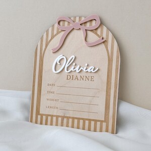 May include: An arched wooden birth announcement sign featuring a light wood grain with vertical stripes. It includes a raised dusty pink acrylic bow at the top, the white 3D script name Olivia, the printed name DIANNE, and lines for DATE, TIME, WEIGHT, and LENGTH.
