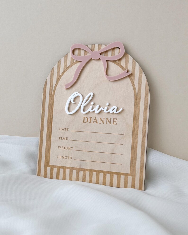 May include: An arched wooden birth announcement sign featuring a light wood grain with vertical stripes. It includes a raised dusty pink acrylic bow at the top, the white 3D script name Olivia, the printed name DIANNE, and lines for DATE, TIME, WEIGHT, and LENGTH.