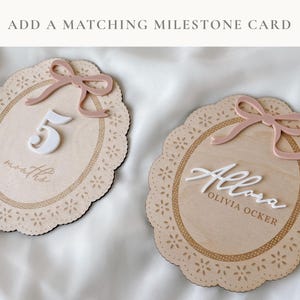 May include: Two oval wooden milestone plaques with scalloped edges and laser-cut floral details. One plaque features a white number 5, the word months, and a dusty pink bow. The other features the white script name Allora, Olivia Ocker, and a bow.