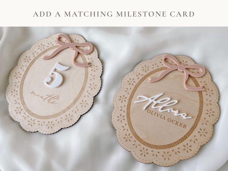 May include: Two oval wooden milestone plaques with scalloped edges and laser-cut floral details. One plaque features a white number 5, the word months, and a dusty pink bow. The other features the white script name Allora, Olivia Ocker, and a bow.
