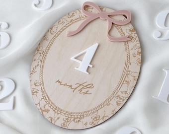 May include: An oval wooden milestone plaque featuring a light wood grain, a white number 4, and the script text months. A dusty pink acrylic bow is attached at the top. The plaque is surrounded by loose white acrylic numbers on a white fabric.