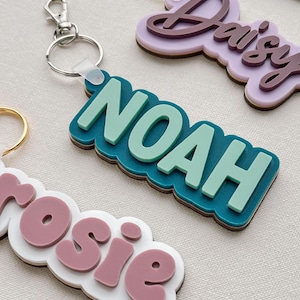 Puede incluir: Primer plano de llaveros personalizados con nombres. El llavero de "Noah" es de color turquesa y verde claro, sujeto a un llavero plateado. Otros llaveros presentan los nombres "Daisy" y "Rosie" en varios colores. Los llaveros tienen un dise&ntilde;o en capas.