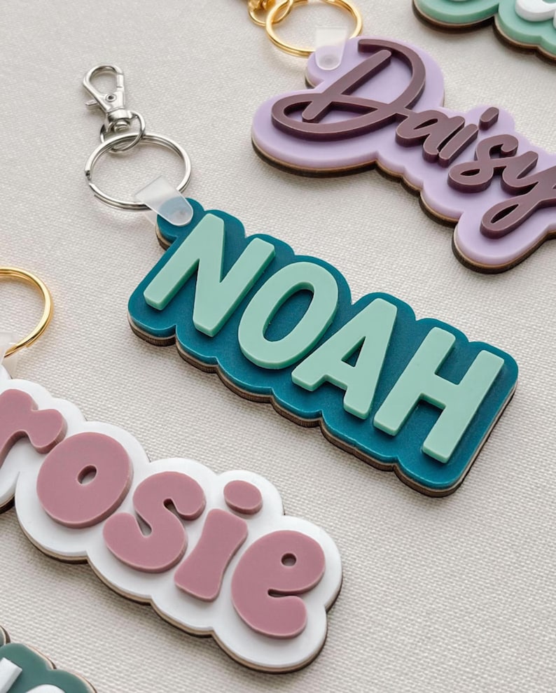 Puede incluir: Primer plano de llaveros personalizados con nombres. El llavero de "Noah" es de color turquesa y verde claro, sujeto a un llavero plateado. Otros llaveros presentan los nombres "Daisy" y "Rosie" en varios colores. Los llaveros tienen un dise&ntilde;o en capas.