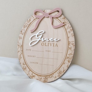 Pu&ograve; includere: Una targa ovale in legno per annuncio di nascita con un fiocco in acrilico rosa polvere sulla parte superiore. La targa mostra il nome Grace OLIVIA in testo bianco e marrone, con linee stampate per data, ora, peso e lunghezza, bordata da fiori.