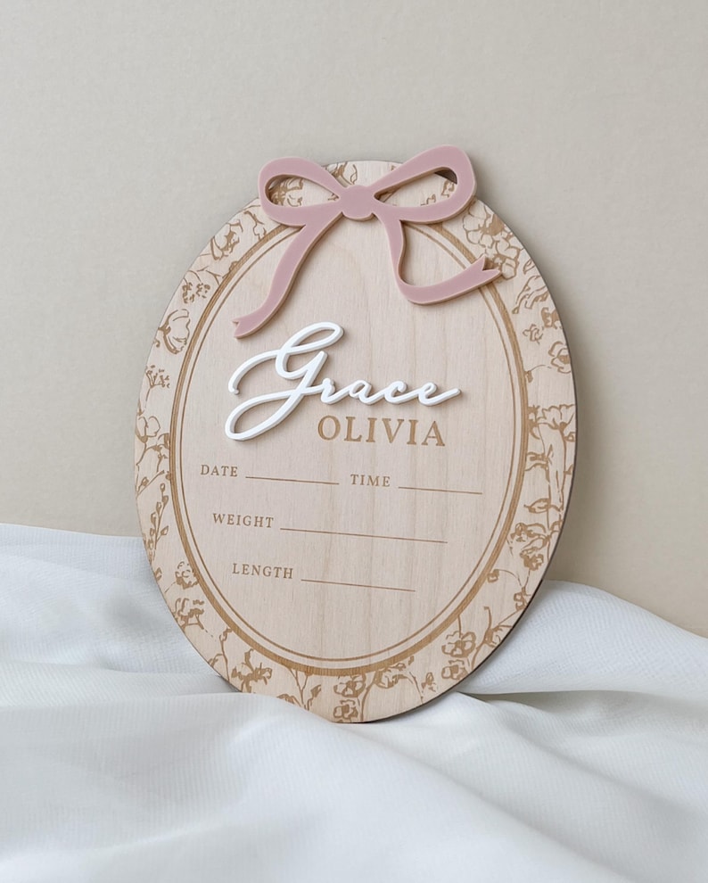 Pu&ograve; includere: Una targa ovale in legno per annuncio di nascita con un fiocco in acrilico rosa polvere sulla parte superiore. La targa mostra il nome Grace OLIVIA in testo bianco e marrone, con linee stampate per data, ora, peso e lunghezza, bordata da fiori.
