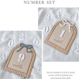 May include: An interchangeable number set featuring two arched wooden plaques with striped borders, one topped with a blue bow and the other with a white bow. The plaques display the text month or months, accompanied by a set of white numbers 0-9.