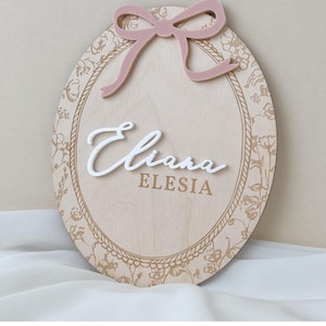Pu&ograve; includere: Una targa ovale in legno con bordo floreale inciso al laser e cornice a corda. La targa riporta il nome Eliana in corsivo bianco in rilievo sopra il nome ELESIA in caratteri dorati, sormontata da un fiocco in acrilico rosa polvere.