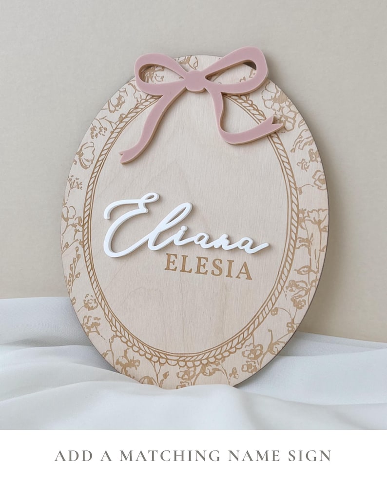 Pu&ograve; includere: Una targa ovale in legno con bordo floreale inciso al laser e cornice a corda. La targa riporta il nome Eliana in corsivo bianco in rilievo sopra il nome ELESIA in caratteri dorati, sormontata da un fiocco in acrilico rosa polvere.