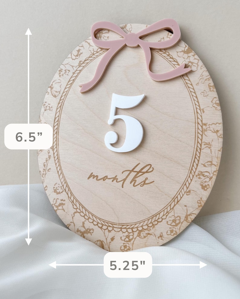 Pu&ograve; includere: Una targa ovale in legno per tappe fondamentali con un fiocco in acrilico rosa polvere sulla parte superiore, il numero 5 in acrilico bianco e la parola months in corsivo. La targa ha un bordo floreale e misura 16,51 cm di altezza e 13,34 cm di larghezza.