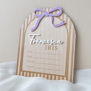 May include: An arched wooden birth announcement sign featuring a light brown and white striped pattern. It includes a lavender acrylic bow at the top, the white script name Francesca, the name IRIS in brown, and lines for date, time, weight, and length.