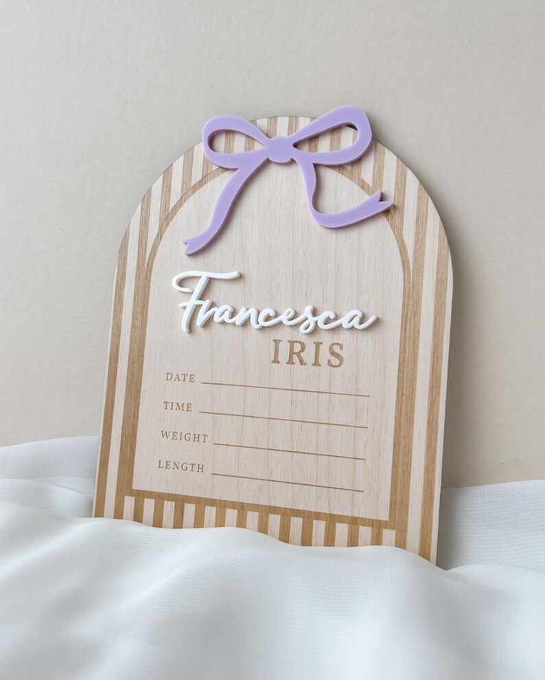 May include: An arched wooden birth announcement sign featuring a light brown and white striped pattern. It includes a lavender acrylic bow at the top, the white script name Francesca, the name IRIS in brown, and lines for date, time, weight, and length.