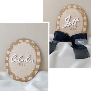 K&ouml;nnte beinhalten: Zwei ovale Holzschilder mit einem hellbraunen Karomuster am Rand. Ein Schild zeigt den wei&szlig;en 3D-Schriftzug Caladin und den braunen Text RHETT. Das andere Schild zeigt den wei&szlig;en 3D-Schriftzug Jett und braunen Text HAYES mit einer marineblauen Schleife.