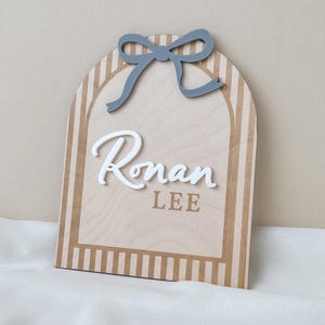May include: An arched wooden sign featuring a light wood grain finish with vertical tan stripes along the border. A grey cutout bow is attached at the top. The name Ronan is displayed in raised white script, with the name LEE in tan block letters.