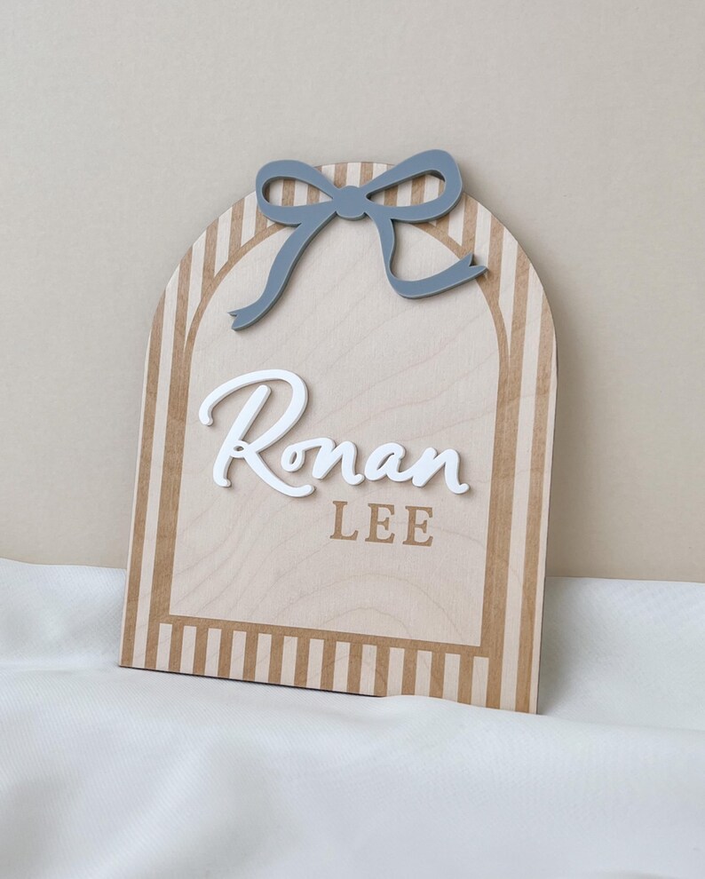 May include: An arched wooden sign featuring a light wood grain finish with vertical tan stripes along the border. A grey cutout bow is attached at the top. The name Ronan is displayed in raised white script, with the name LEE in tan block letters.