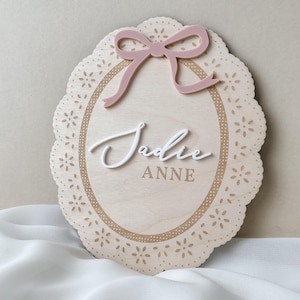 May include: An oval wooden plaque with a scalloped edge featuring a laser-cut floral pattern. The plaque displays a dusty pink acrylic bow at the top and the name "Jadie" in raised white script lettering above the printed text "ANNE" in brown.