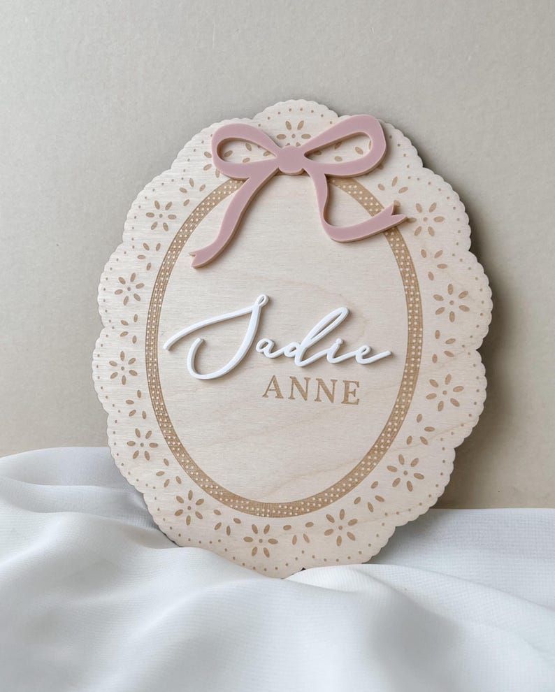 May include: An oval wooden plaque with a scalloped edge featuring a laser-cut floral pattern. The plaque displays a dusty pink acrylic bow at the top and the name "Jadie" in raised white script lettering above the printed text "ANNE" in brown.