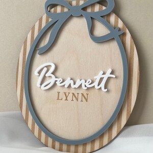 Puede incluir: Letrero ovalado de madera con lazo y borde grises. El letrero presenta el nombre "Bennett" en escritura blanca sobre el nombre "LYNN" en una fuente m&aacute;s peque&ntilde;a. El fondo es de madera clara con rayas verticales.