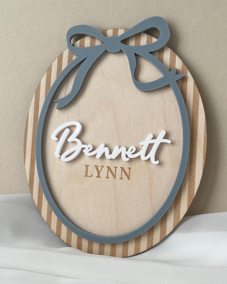 Puede incluir: Letrero ovalado de madera con lazo y borde grises. El letrero presenta el nombre "Bennett" en escritura blanca sobre el nombre "LYNN" en una fuente m&aacute;s peque&ntilde;a. El fondo es de madera clara con rayas verticales.