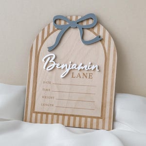May include: An arched wooden birth announcement sign featuring a light wood grain with vertical tan stripes. A raised blue bow sits at the top. The name Benjamin Lane is in white and tan lettering, with blank lines for date, time, weight, and length.