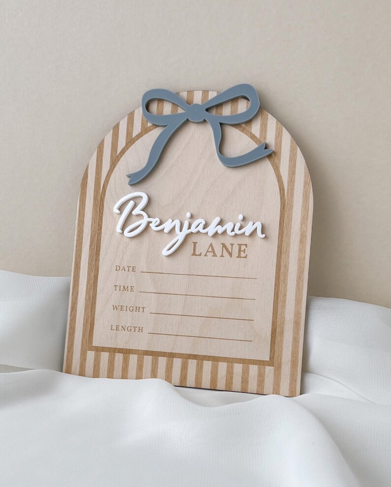 May include: An arched wooden birth announcement sign featuring a light wood grain with vertical tan stripes. A raised blue bow sits at the top. The name Benjamin Lane is in white and tan lettering, with blank lines for date, time, weight, and length.