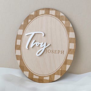 K&ouml;nnte beinhalten: Ein ovales Holzschild mit einem hellbraunen und beigen Karomuster am Rand. In der Mitte steht der Name Troy in erhabener, wei&szlig;er Schreibschrift, w&auml;hrend der Name JOSEPH rechts in einer einfachen, serifenlosen Gro&szlig;schrift gedruckt ist.