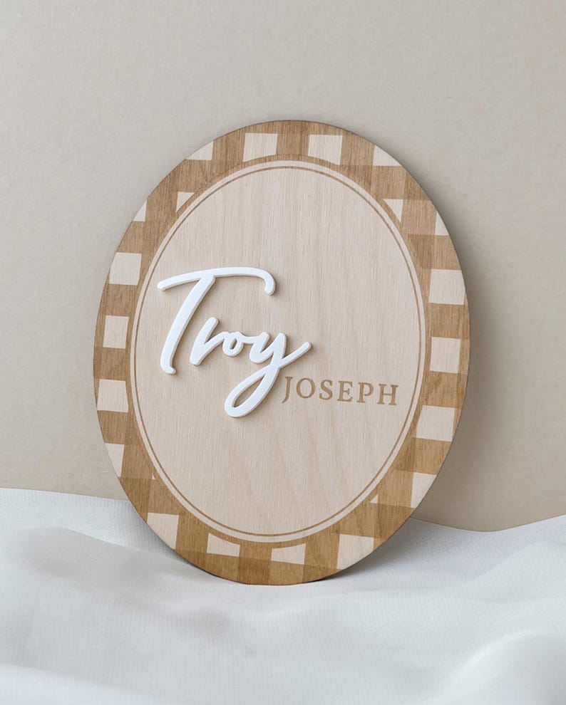 K&ouml;nnte beinhalten: Ein ovales Holzschild mit einem hellbraunen und beigen Karomuster am Rand. In der Mitte steht der Name Troy in erhabener, wei&szlig;er Schreibschrift, w&auml;hrend der Name JOSEPH rechts in einer einfachen, serifenlosen Gro&szlig;schrift gedruckt ist.