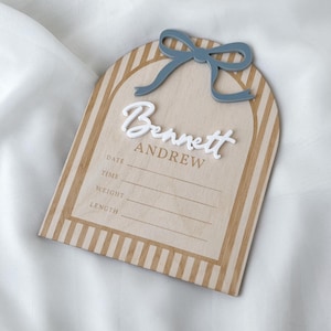 May include: An arched wooden birth announcement sign featuring a light wood finish with tan vertical stripes. The sign displays the name Bennett Andrew in white and tan lettering, a blue bow accent, and lines for date, time, weight, and length.