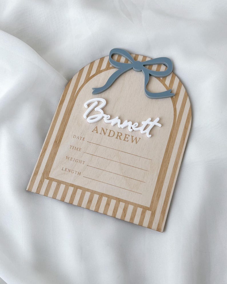 May include: An arched wooden birth announcement sign featuring a light wood finish with tan vertical stripes. The sign displays the name Bennett Andrew in white and tan lettering, a blue bow accent, and lines for date, time, weight, and length.