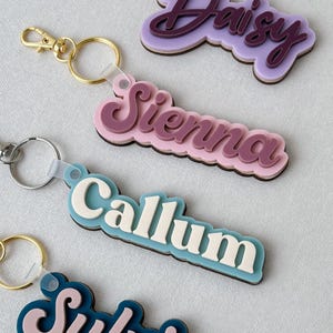 May include: Four layered acrylic keychains featuring the names Daisy, Sienna, Callum, and Sylvia in various color combinations. Each keychain has a metal ring attached by a translucent plastic loop, showcasing stacked lettering with a dark base layer.