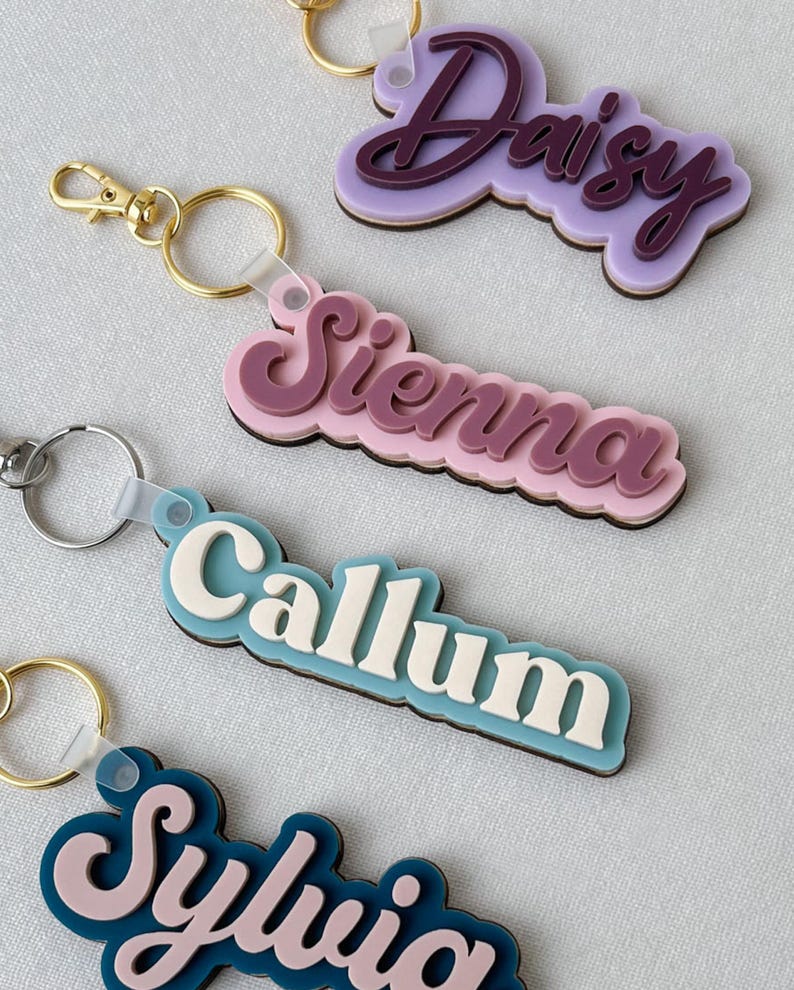 May include: Four layered acrylic keychains featuring the names Daisy, Sienna, Callum, and Sylvia in various color combinations. Each keychain has a metal ring attached by a translucent plastic loop, showcasing stacked lettering with a dark base layer.