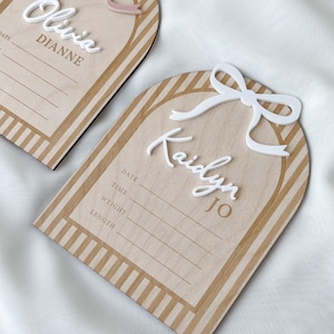 May include: Two arch-shaped wooden birth announcement signs with striped borders. One features a dusty pink bow and the name Olivia Dianne, the other a white bow and the name Kaidyn Jo. Both include printed lines for date, time, weight, and length.