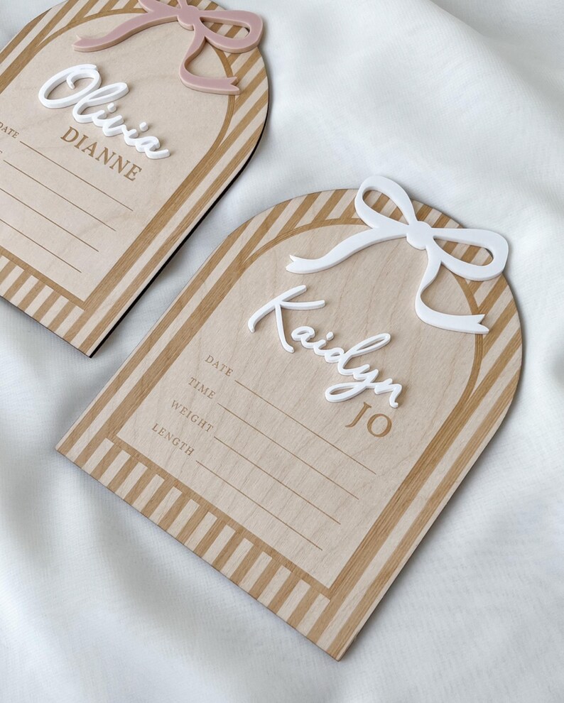 May include: Two arch-shaped wooden birth announcement signs with striped borders. One features a dusty pink bow and the name Olivia Dianne, the other a white bow and the name Kaidyn Jo. Both include printed lines for date, time, weight, and length.