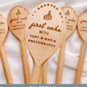 K&ouml;nnte beinhalten: Ein heller Holzl&ouml;ffel mit einer Lasergravur eines Kuchensymbols und dem Text I SMASHED MY first cake WITH TONY & MARIA PHOTOGRAPHY. Der L&ouml;ffel hat eine glatte Oberfl&auml;che und eine abgerundete Form und wird in einer Gruppe &auml;hnlicher Artikel gezeigt.