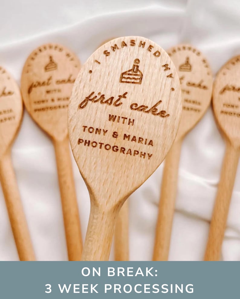 K&ouml;nnte beinhalten: Ein heller Holzl&ouml;ffel mit einer Lasergravur eines Kuchensymbols und dem Text I SMASHED MY first cake WITH TONY & MARIA PHOTOGRAPHY. Der L&ouml;ffel hat eine glatte Oberfl&auml;che und eine abgerundete Form und wird in einer Gruppe &auml;hnlicher Artikel gezeigt.