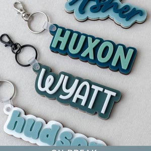 May include: Four custom keychains featuring layered acrylic name tags in shades of blue, teal, and white. Each keychain includes a metal ring and a lobster clasp. The names Asher, HUXON, WYATT, and hudson are displayed in various fonts.