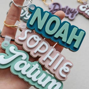 Puede incluir: Llaveros con nombres como "Noah", "Sophie" y "Tatum". Los llaveros tienen un dise&ntilde;o en capas con una combinaci&oacute;n de colores turquesa, rosa y blanco. Cada llavero incluye un anillo met&aacute;lico.