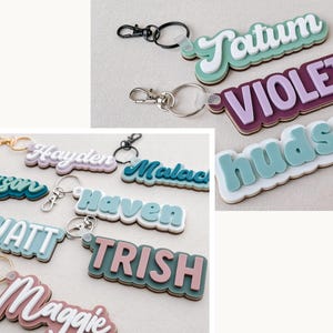 Puede incluir: Una colecci&oacute;n de llaveros personalizados con nombres variados. Los llaveros presentan nombres como "Tatum", "Violet" y "Hudson" en un dise&ntilde;o en capas con efecto 3D. Cada llavero tiene un cierre met&aacute;lico.